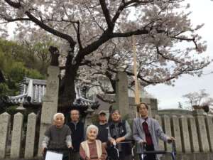 施設の入居者と職員が桜の木の下で集合写真を撮影している様子